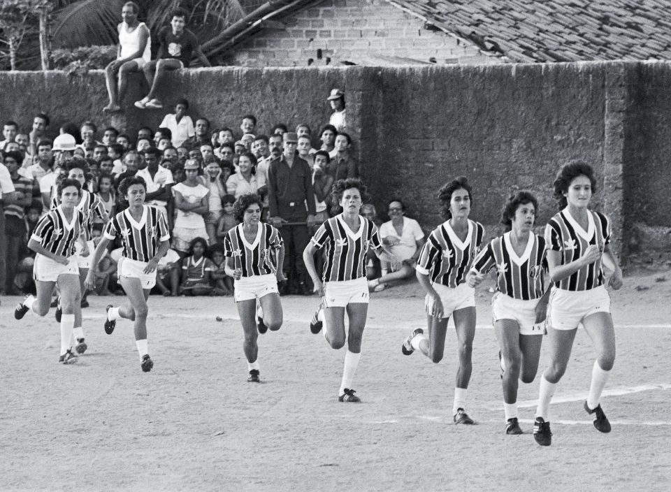 Imagem em preto e branco de mulheres vestidas com uniforme de futebol e torcedores ao fundo