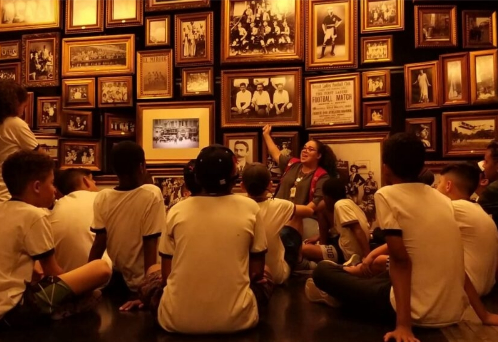 Imagem de um grupo de crianças e uma educadora do museu em uma sala com quadros