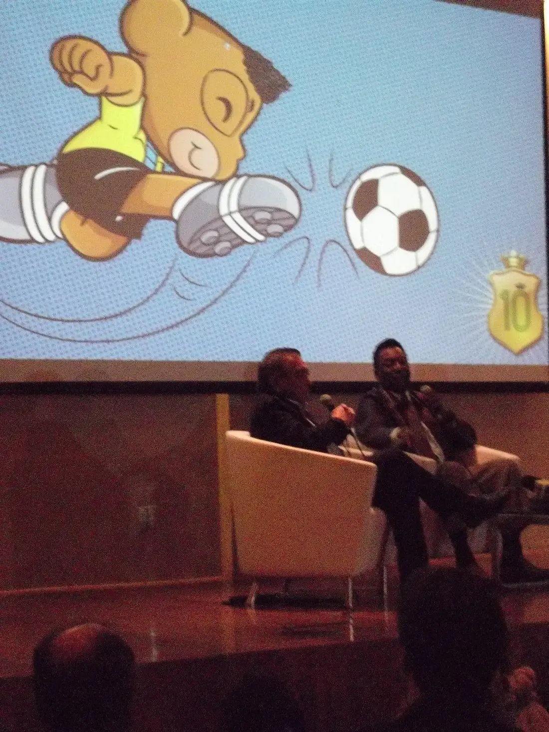 Pelé sentado no palco do auditório do Museu do Futebol. Ao fundo o telão projeta uma imagem do personagem Pelezinho, de Maurício de Souza.
