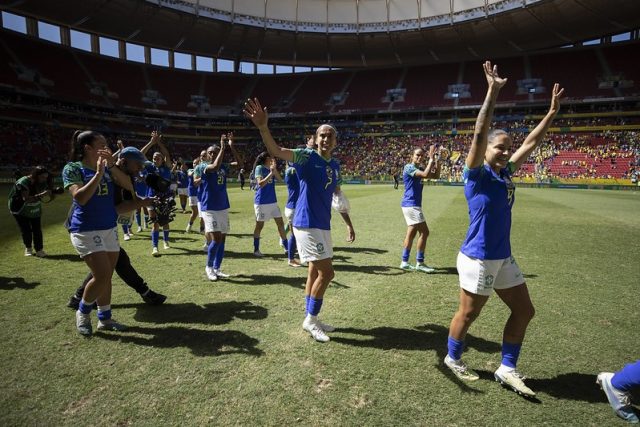 Seleção feminina deixa o campo após uma partida, vestindo os uniformes azuis. Elas acenam para o público.
