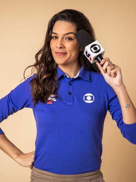 Renata Silveira veste o uniforme da Globo e SportV, que é uma camisa azul escura tipo polo de mangas compridas. Ela tem cabelo escuro, longo, e segura o microfone perto do rosto.