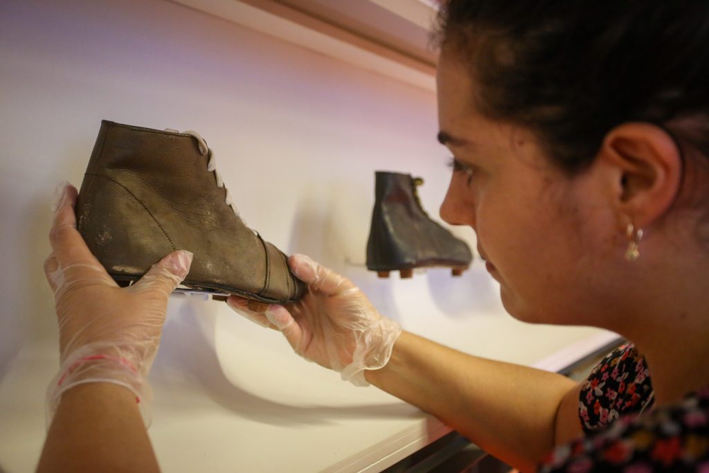 Foto de uma museóloga retirando uma chuteira antiga de couro de uma vitrine. Ela usa luvas.