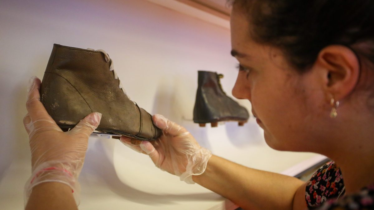 Foto de uma museóloga retirando uma chuteira antiga de couro de uma vitrine. Ela usa luvas.