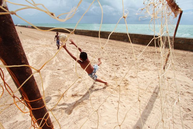 Duas crianças jogam boa em uma praia, uma delas tenta defender o gol contra o chute da outra. A traves estão enferrujadas e a rede puída. A cena é fotogafada através das redes.