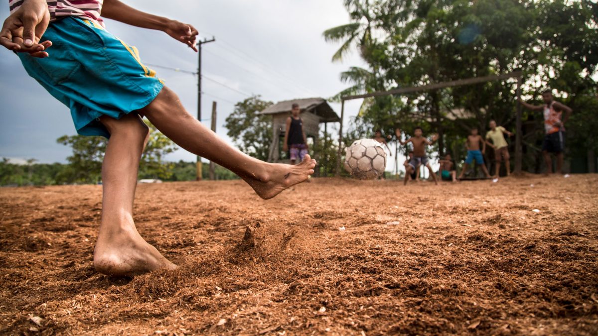 Um menino de shorts azuis chuta uma bola de futebol em direção a outras crianças. Ela está descalça e brinca sobre um chão de terra, em um lugar rural.