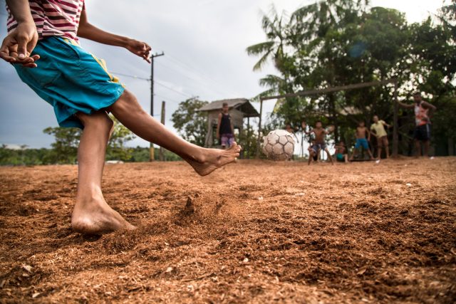 Um menino de shorts azuis chuta uma bola de futebol em direção a outras crianças. Ela está descalça e brinca sobre um chão de terra, em um lugar rural.