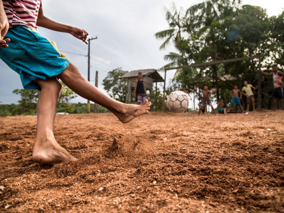 Um menino de shorts azuis chuta uma bola de futebol em direção a outras crianças. Ela está descalça e brinca sobre um chão de terra, em um lugar rural.