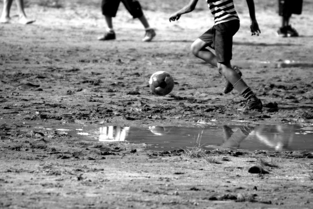 Imagem em preto e branco destaca os pés de uma criança jogando em um terrão, com bastante lama.