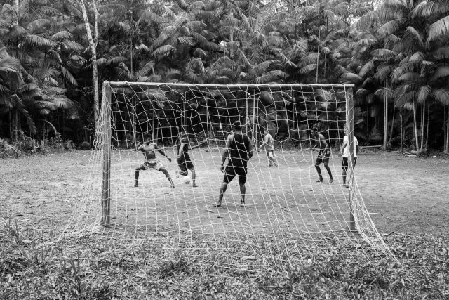 Imagem em preto e branco vista por trás de um gol. Pessoas jogam em um campo de grama; atrás do campo se veem palmeiras.