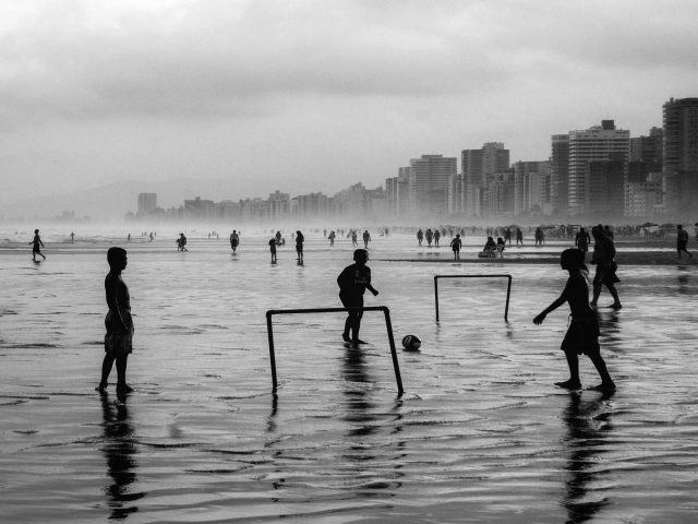 Imagem em preto e branco com cena de praia: um grupo de meninos joga bola na água rasa, com duas barrinhas improvisadas.