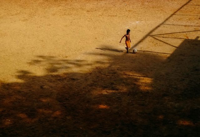 Uma criança solitária brinca com uma bola em um campo de terra. Apenas ela aparece na cena.