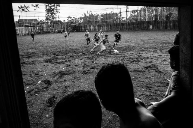 Imagem em preto e branco de crianças brincando de futebol em um campo de terra. Em primeiro plano se vê as silhuetas de outras crianças que assistem ao jogo da janela.