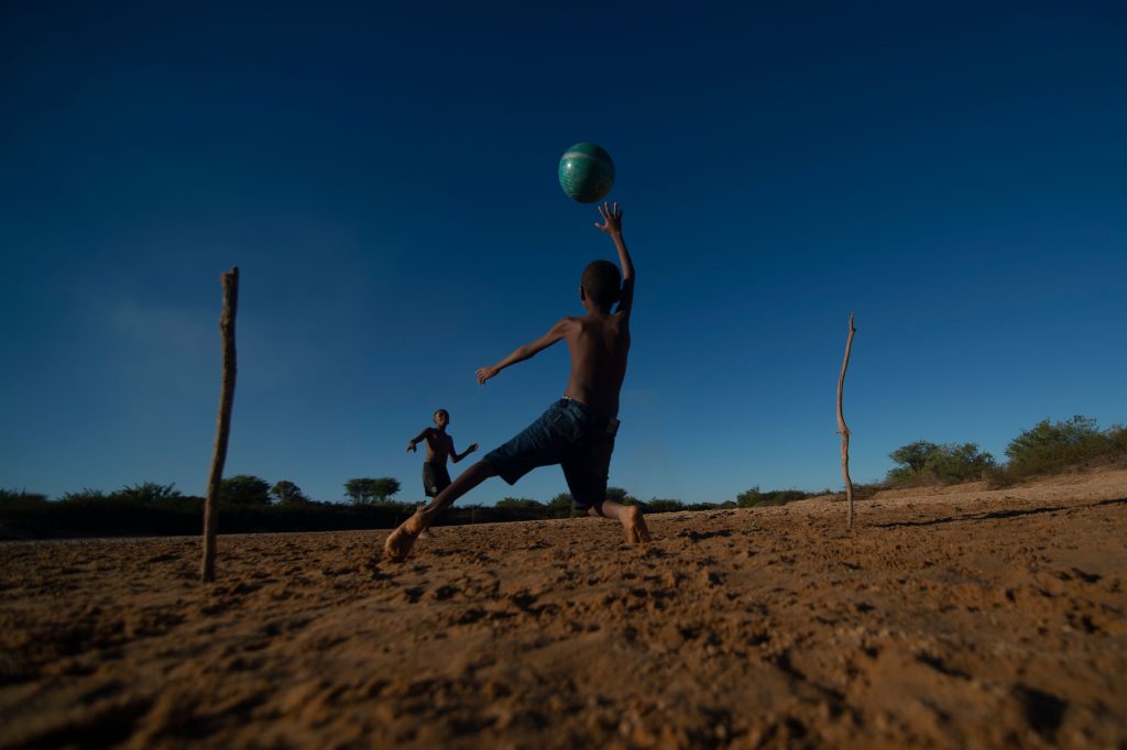 Crianças brincam de futebol em um campo de terra com traves improvisadas de galhos de árvores. Chama a atenção o céu muito azul, sem nuvens.
