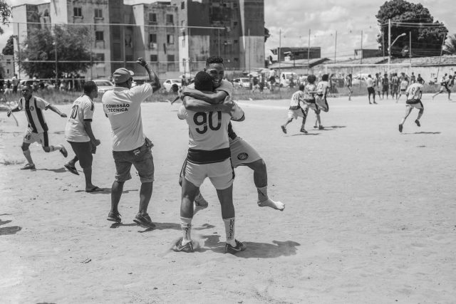 Imagem em preto e branco de homens comemorando um gol em um campo de areia. Um condomínio aparece ao fundo.