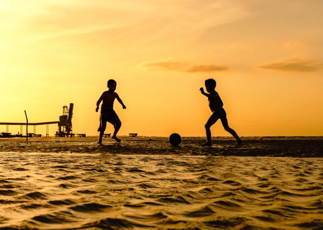 Duas crianças brincam de jogar bola na praia ao pôr do sol. A imagem está em contra luz.