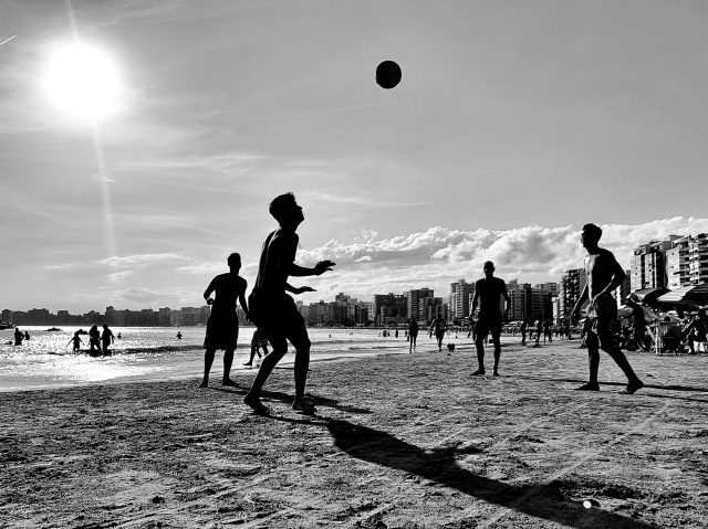 Homens jogam altinha em uma praia. Imagem em preto e branco.