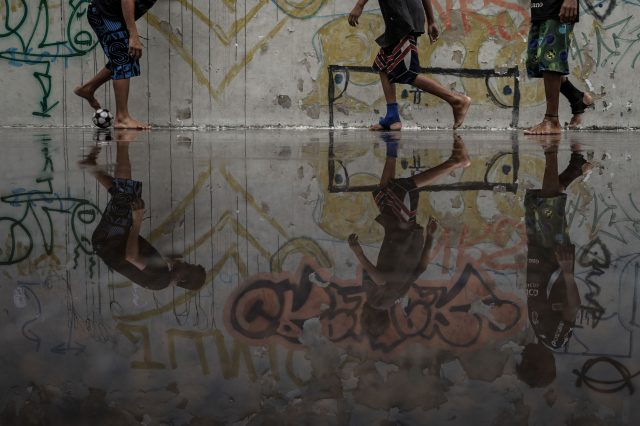 A imagem mostra o reflexo em uma grande poça d'água de três jovens brincando de futebol em um ambiente urbano. O gol está pintado na parede, que é toda grafitada.