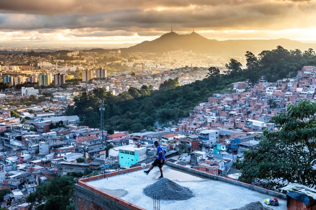 Um homem faz embaixadinhas em uma laje na periferia de São Paulo. Ao fundo se vê uma favela, prédios à distância e o Pico do Jaraguá.