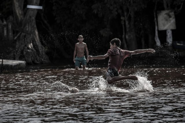 Crianças brincam de jogar bola na água, no que parece ser um rio ou lago. Destaca-se na imagem a cortina de gotas quando uma delas chuta a bola.