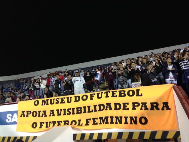 Faixa amarela na arquibancada de um estádio com os dizeres "O Museu do Futebol apoia a Visibilidade para o Futebol Feminino"