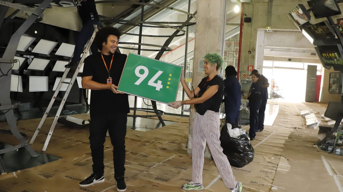 Um funcionário e uma funcionária do Museu do Futebol carregam uma placa verde com 94 escrito e quatro estrelas douradas,