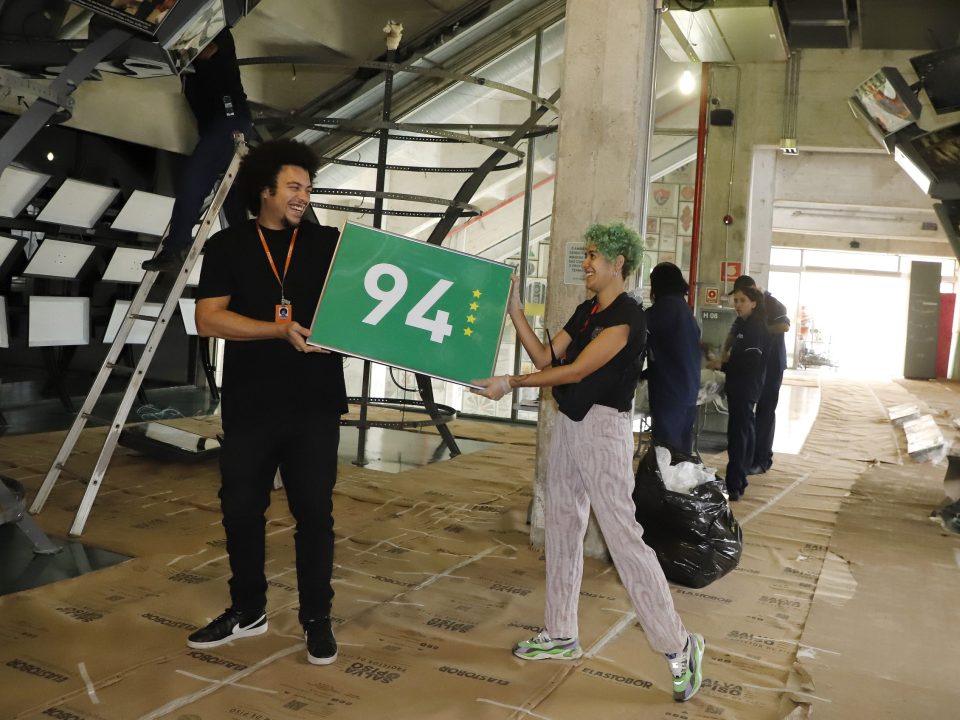 Um funcionário e uma funcionária do Museu do Futebol carregam uma placa verde com 94 escrito e quatro estrelas douradas,