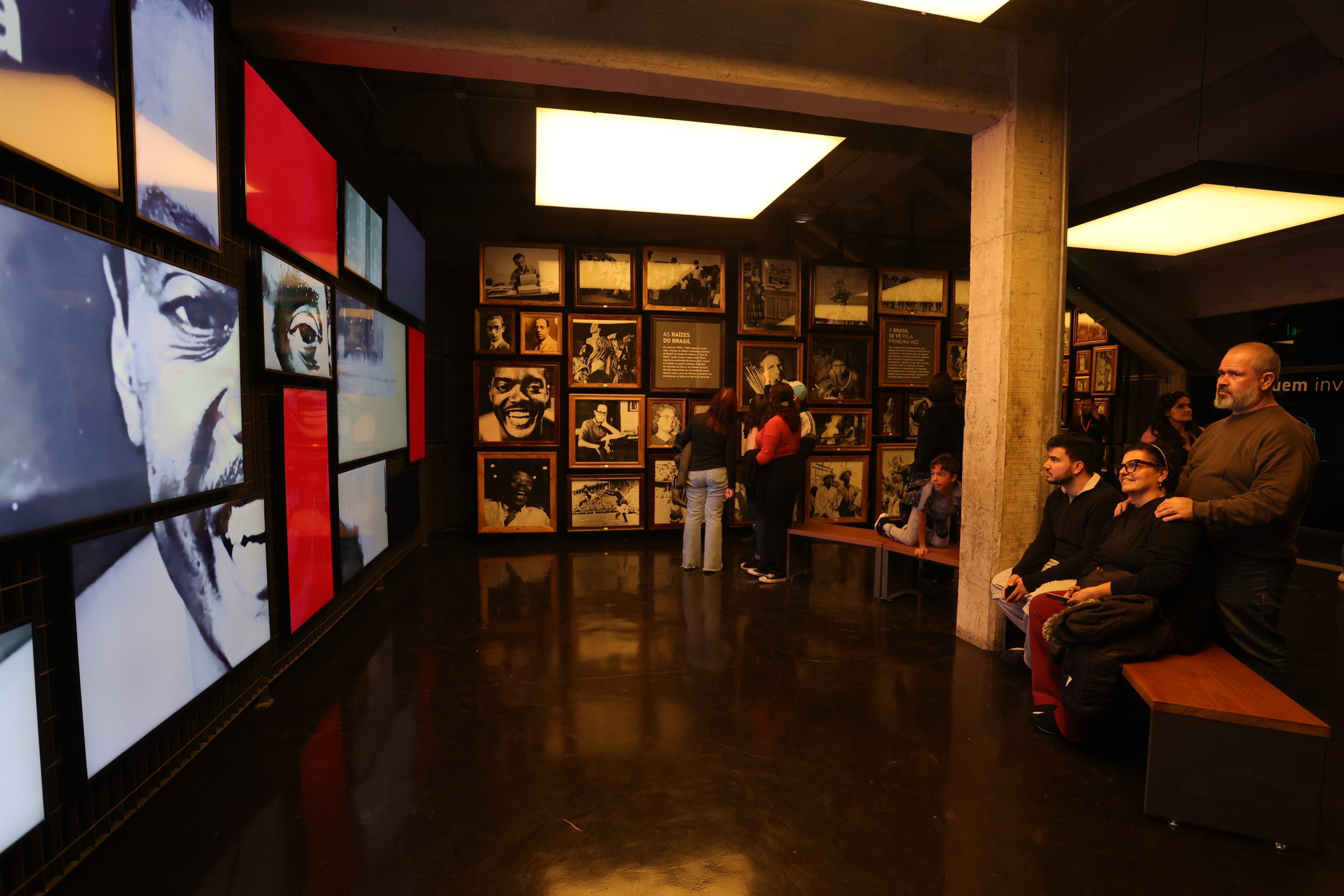 Pessoas assistem à obra audiovisual da Sala, composta por imagens que aparecem em várias telas de TV, dispostas como se fossem um quebra-cabeças