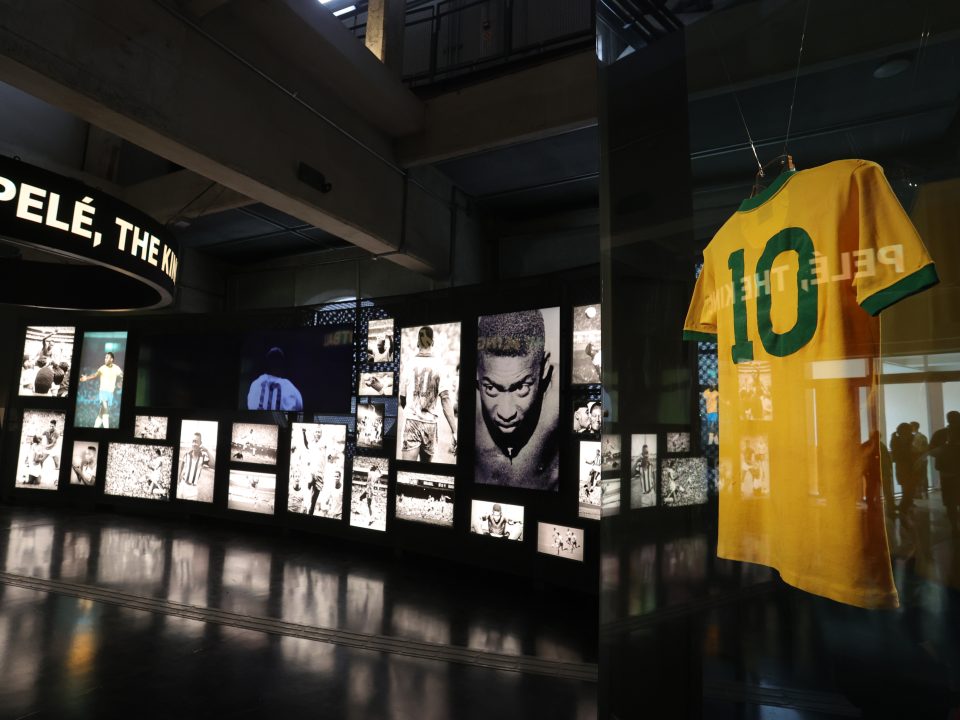 Em primeiro plano aparece a camisa 10 amarela usada por Pelé na Copa de 1970. No fundo há um painel com várias imagens iluminadas, fotos em preto e branco do Rei.