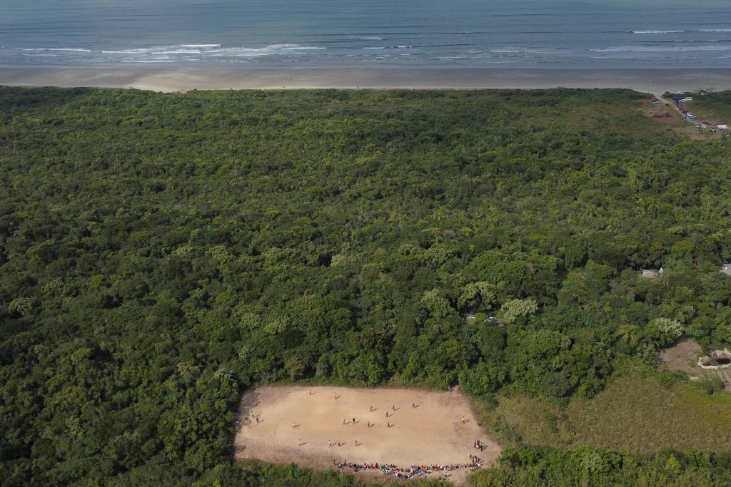 Imagem registrada do alto e três diferentes cenários a compõe: o oceano, a floresta e o campo no mio da floresta com pessoas jogando futebol.