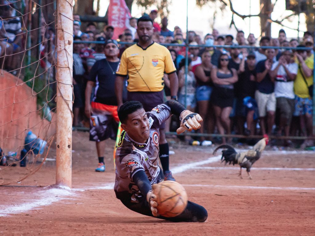 Na imagem um cenário interessante: o goleiro se esforça para defender a bola. Atento o bandeirinha olha a defesa. Torcedores aglomerados ao alambrado assistem o lance e enquanto isso, um galo circula tranquilamente no campo.