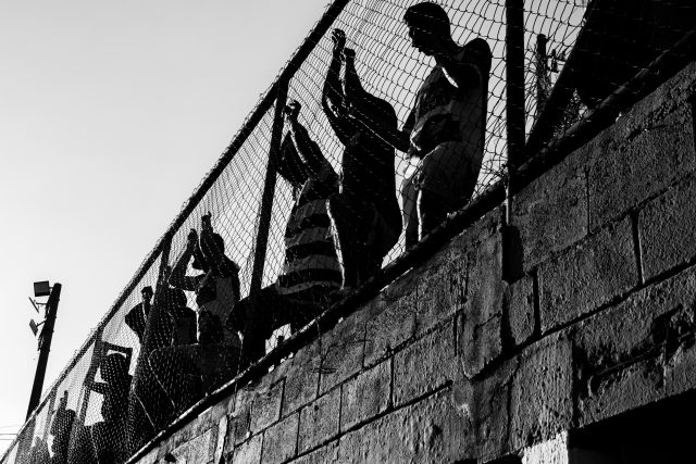 Imagem em preto e branco registra torcedores grudados ao alambrado assistindo à partida de futebol.