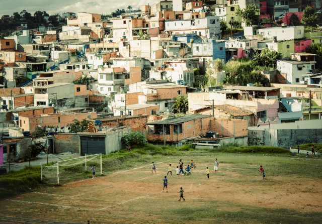 Ao redor de uma favela, em uma imagem registrada do alto, crianças jogam bola em um campo de futebol.