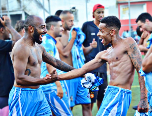 Dois homens sem camisa e de bermuda azul se cumprimentam felizes, comemorando uma possível vitória após o jogo.