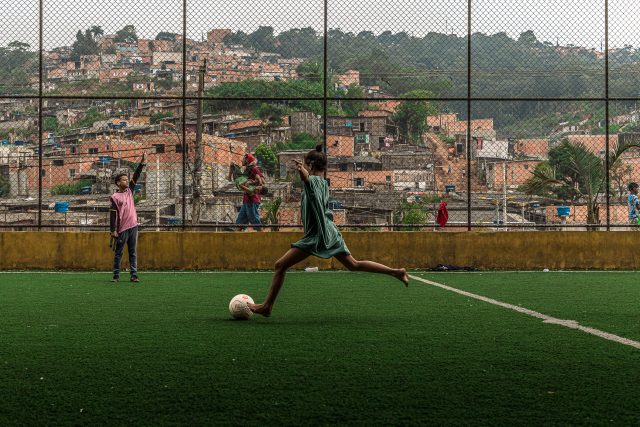 Uma criança descalça chuta uma bola em um campo de grama sintética. Ao fundo, casas de periferia compõem o cenário.