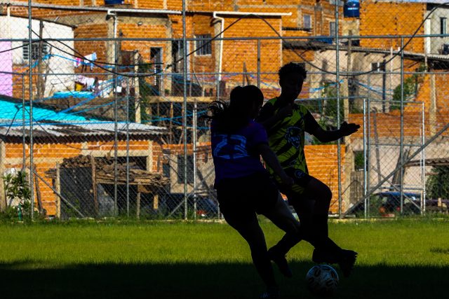 A silhueta de duas mulheres marca a disputa da bola. O campo é de grama e, ao fundo, casas de tijolo e sem pintura compõem o cenário.