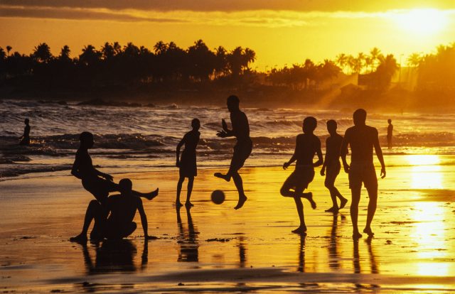 O pôr-do-sol, os últimos raios do dia contemplam os jovens batendo uma bolinha à beira do mar.