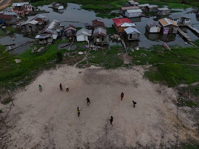 Imagem registrada do alto. Casas de pau-a-pique compõem o improvisado campo de futebol de areia. Jovens jogam futebol nele.