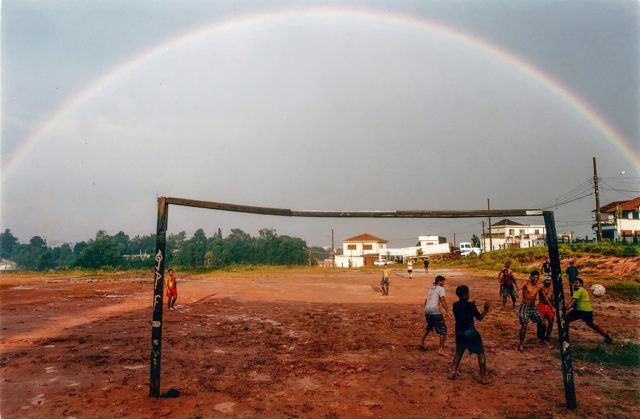 Um arco-íris compõe o cenário de crianças jogando futebol em um campo cheio de lama. A trave é improvisada e as crianças observam o destino da bola.