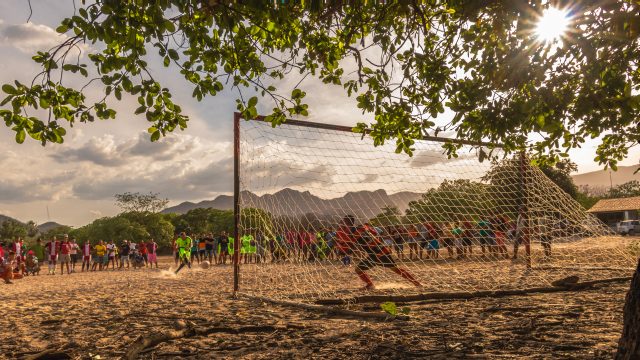 Em um campo lotado torcedores e jogadores assistem ao atleta batendo um pênalti. O campo é de areia e iluminado pelo sol.