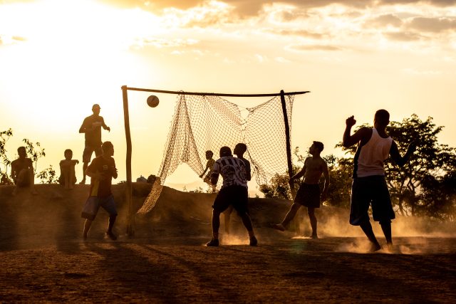 Imagem de pessoas em uma partida de futebol. A bola entra na rede improvisada.