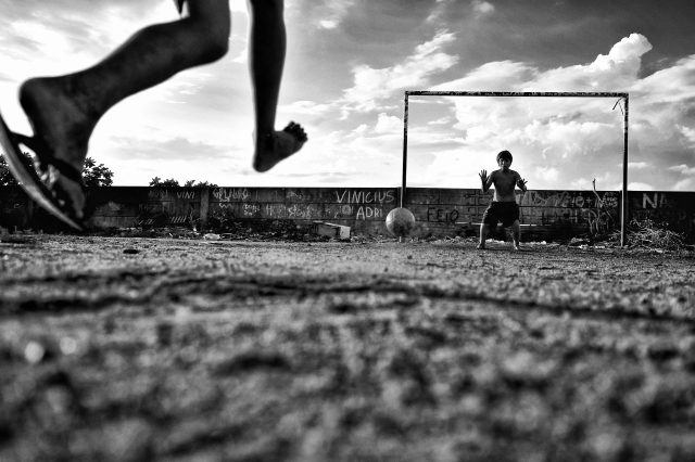 Imagem em preto e branco e um interessante cenário. Os pés de uma criança (com um pé descalço e o outro com chinelo) faz o chute ao gol. Do outro lado, uma criança sem camisa se prepara para uma possível defesa.