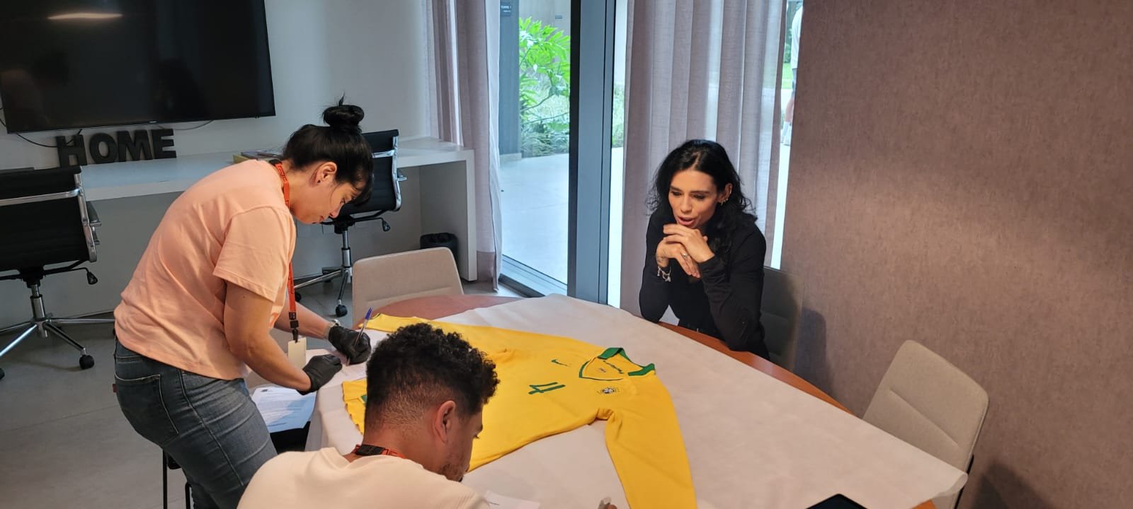 Três pessoas posicionadas ao redor de uma mesa oval, revestida por TNT, situada em um ambiente fechado. Estendida sobre a mesa, há uma camisa esportiva nas cores verde e amarela. Três pessoas estão na imagem. À esquerda, uma mulher em pé e curvada sobre a mesa toma notas em um papel; abaixo, um homem sentado também faz registros; e, do lado oposto, outra mulher sentada, com os cotovelos apoiados sobre a mesa e as mãos abaixo do queixo, apresenta movimentos labiais.
