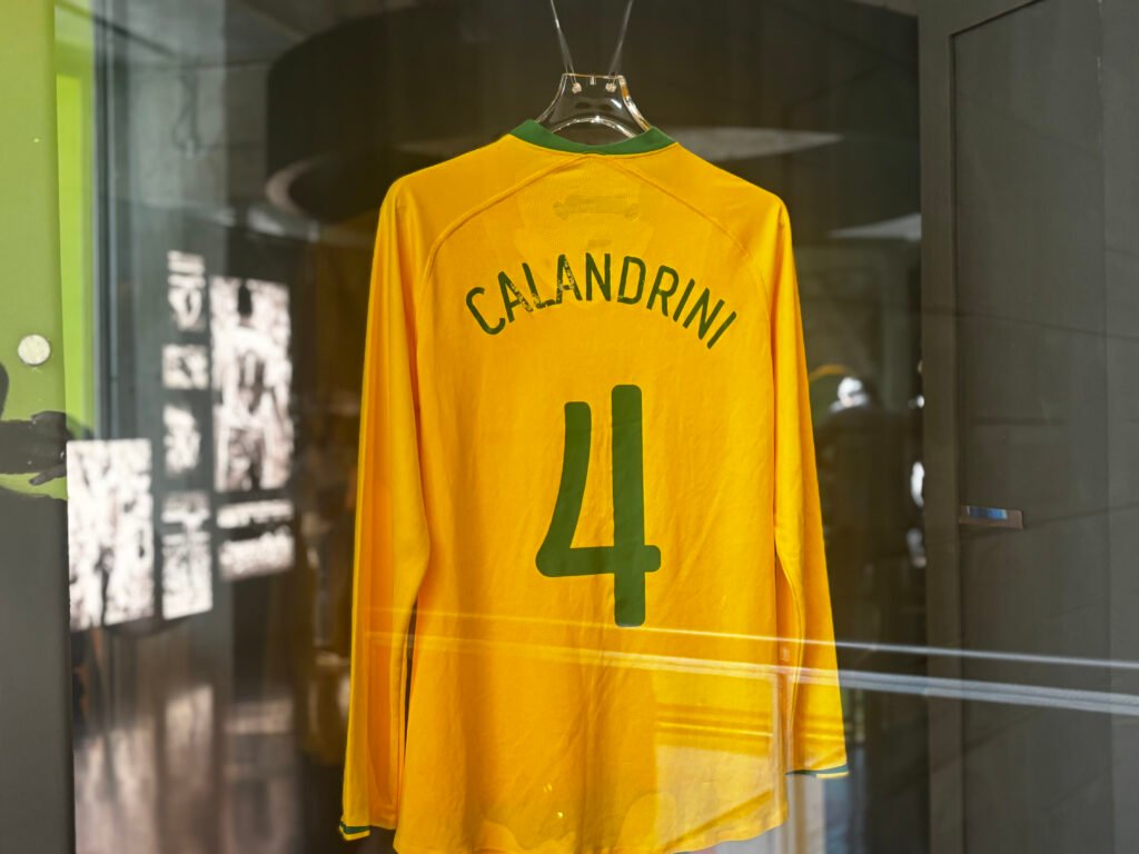 Camisa esportiva nas cores verde e amarela, pendurada por cabos de aço em cabide de acrílico, instalada dentro de vitrine vertical. Nela, constam o nome da atleta e o número de sua camisa: Calandrini, 4.