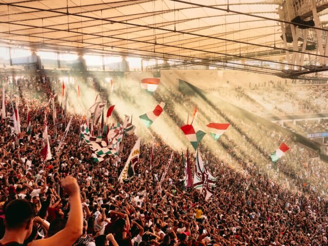 162_FOTO_YgorMartins Em um estádio lotado, vê-se uma torcida animada agitando bandeiras nas cores vermelho, branco e verde. Uma faixa de lux amarela passa pela cobertura do estádio e ilumina o momento.
