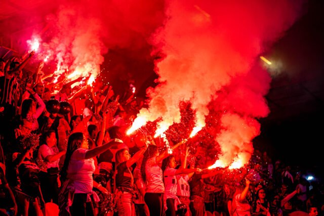 25_FelipeGouveia_kokfotos-KokFotos Uma arquibancada se ilumina de vermelho com sinalizadores. Em primeiro plano, nota-se que há muitas torcedoras mulheres.