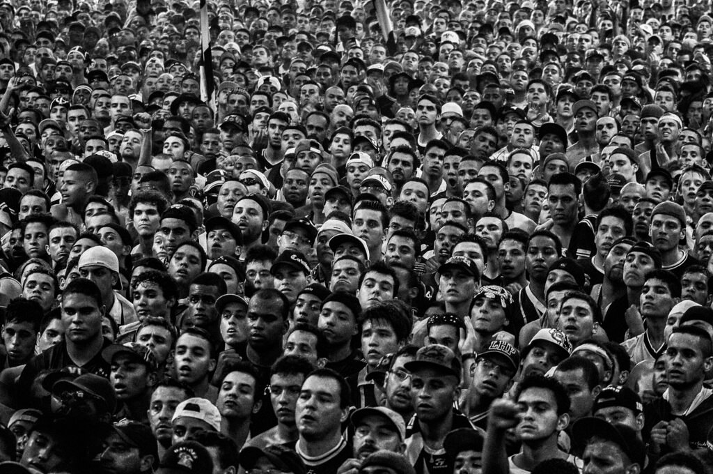 Foto em preto e branco mostrando os rostos de centenas de torcedores do Corinthians acompanhando um jogo na quadra da Gaviões da Fiel. Não se vê a tela, apenas as pessoas.