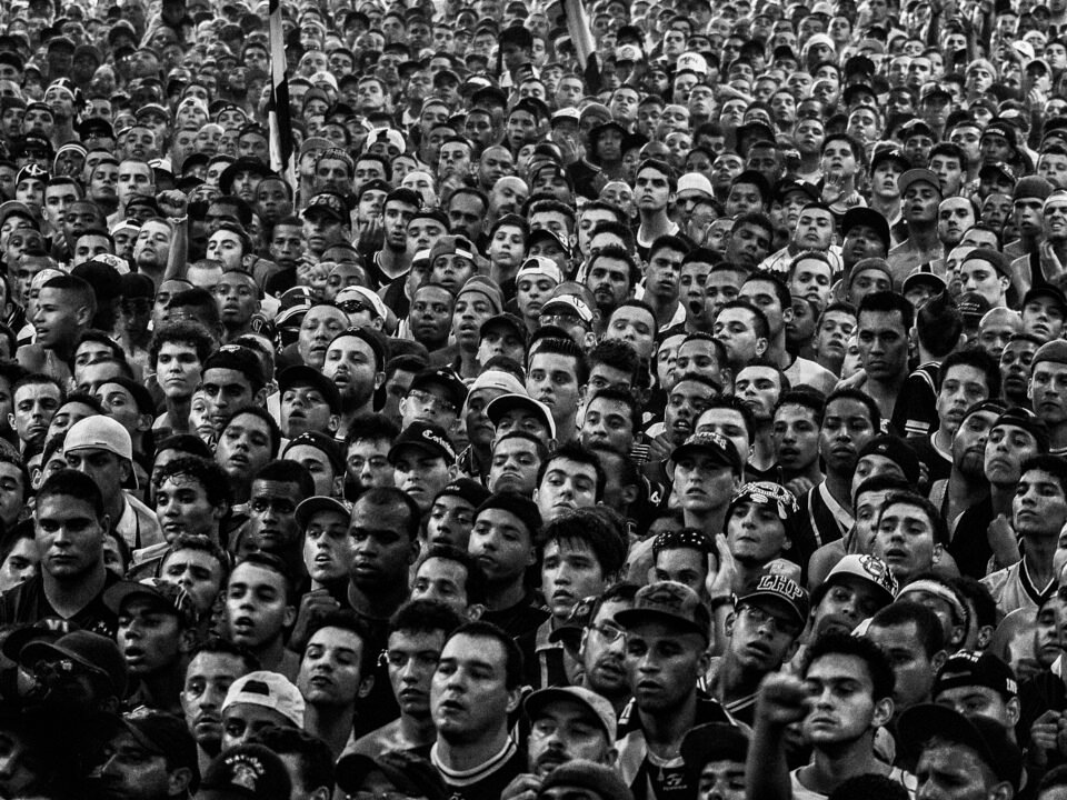 Foto em preto e branco mostrando os rostos de centenas de torcedores do Corinthians acompanhando um jogo na quadra da Gaviões da Fiel. Não se vê a tela, apenas as pessoas.
