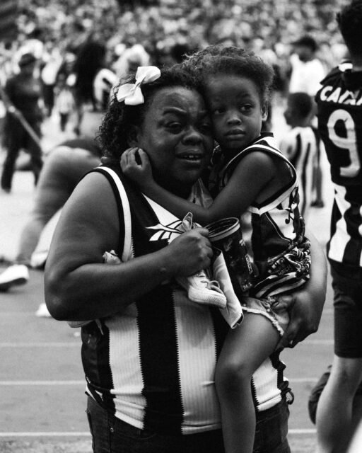 64_FOTO_JoanaBrandao Uma mulher negra com uma camisa de listras preto e brancas, com uma grande flor branca no cabelo, tem uma criança também negra no colo. As duas estão emocionadas.