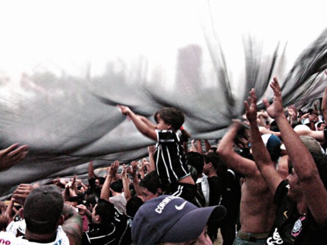 86_ArthurGomesCosta_62,51x45,88_300DPI_ Torcida sob um bandeirão preto estendido. Em destaque, uma meinina nas costas de um adulto fica mais alta do que todos.