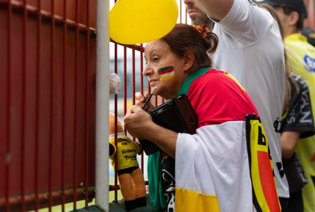 90_FOTO_LuanaGums Uma senhora se agarra à grade que separa a torcida do campo. Ela tem o rosto pintado e está enrolada com uma bandeira do clube. Numa das mãos ela tem um mascote de pelúcia.